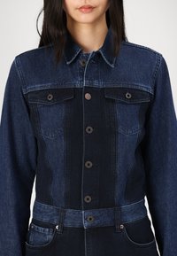 Denim kombinezon z lahkega do temnega gradienta, z gumbastim zapiranjem, dvema žepoma na prsih in pasom s pasom. Teksturen, strukturiran dizajn.