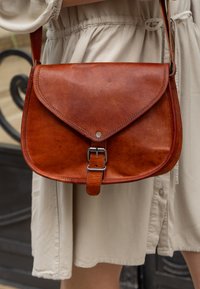 Smeđi kožni crossbody ranac s zakrivljenim preklopom, srebrnim kopčama i glatkom teksturom. Remen je podesiv radi praktičnosti.