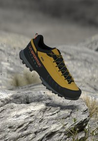 Sapatilha de trail La Sportiva amarela e preta com sola resistente a saltar sobre terreno rochoso com pequenas plantas e partículas de terra.