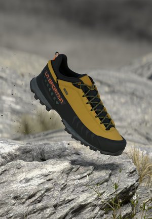 Zapatilla de trail La Sportiva amarilla y negra con suela Rugosa saltando sobre terreno rocoso con pequeñas plantas y partículas de tierra.
