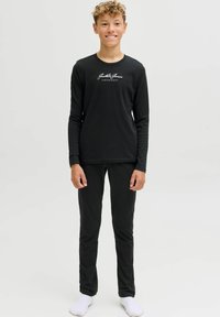 Pusaudzis ar cirtainiem matiem, ģērbies melnā garo piedurkņu Jack & Jones kreklā, melnās biksēs un baltās zeķēs, stāv uz baltas fona.