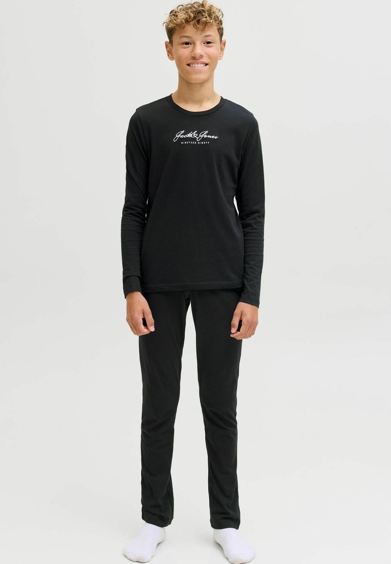 Pusaudzis ar cirtainiem matiem, ģērbies melnā garo piedurkņu Jack & Jones kreklā, melnās biksēs un baltās zeķēs, stāv uz baltas fona.