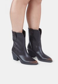 Botine de cowboy din piele maro cu vârf ascuțit, cusături definite și toc înclinat. Dispun de bucle de tragere pentru o încălțare ușoară și o finisare netedă.