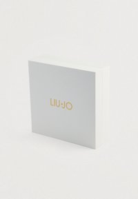 Valkoinen suorakulmainen laatikko, jossa on teksturoitu pinta, etupuolella kultainen 'LIU JO' -logo. Kulmat ovat neliömäiset ja muotoilu on minimalistinen.