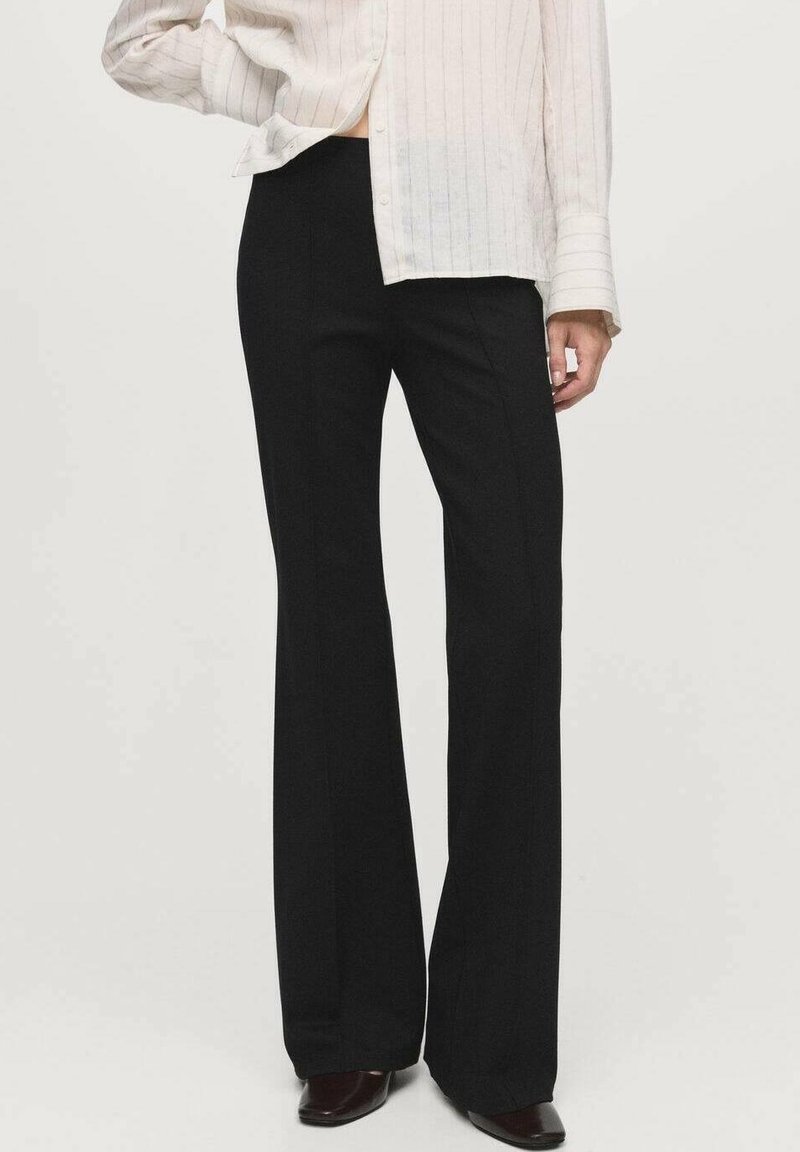 Femme portant un pantalon noir évasé taille haute, une chemise blanche à manches longues et des chaussures pointues foncées, debout devant un fond uni.