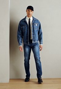 Jeansjakke med to frontlommer, iført over en hvit skjorte og svart slips, kombinert med mørke jeans og svarte sko, alt satt mot en nøytral bakgrunn.