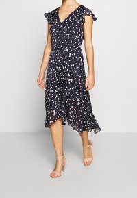 Navy blue dress with pink polka dots, short ruffled sleeves, V-neck, belted waist, and a flared, ruffled hem. Paired with nude heels.