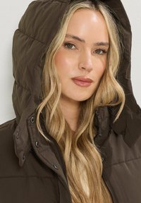 Brun pufferjacka med huva, som har dragkedja och tryckknappar. Yttermaterialet är slätt och vadderat, vilket ger värme och komfort.