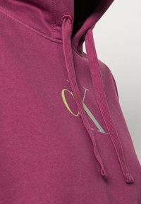 Maroon hoodie with a drawstring hood and front pocket. Features a light green logo on the chest. Made from a soft, textured fabric.