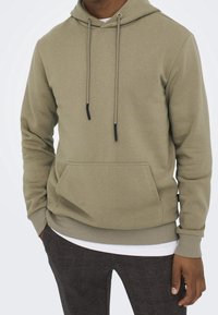 Oliwkowy hoodie wykonany z miękkiego materiału, z przednią kieszenią, sznurkami z czarnymi końcówkami oraz ściągaczami na rękawach i w talii.