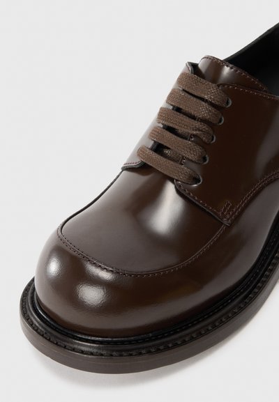 Scarpa di pelle marrone con finitura lucida. Presenta sei lacci marroni, una punta arrotondata e una suola nera a contrasto con dettagli di cucitura.