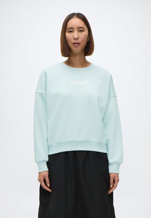 Felpa azzurra chiaro con collo rotondo, spalle cadenti e grafica bianca "XOXO" sul petto. Il tessuto appare morbido e rilassato.