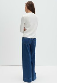 Frau mit langen braunen Haaren, steht mit dem Rücken zur Kamera, trägt ein weißes Langarmshirt und weite blaue Jeans, vor einem einfarbigen Hintergrund.