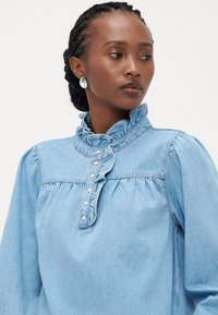 Ανοιχτό μπλε τζιν πουκάμισο με ρυπώσεις στον γιακά, κουμπιά μπροστά και συγκεντρωμένες λεπτομέρειες στους ώμους και το στήθος. Διαθέτει μακρύ μανίκι.