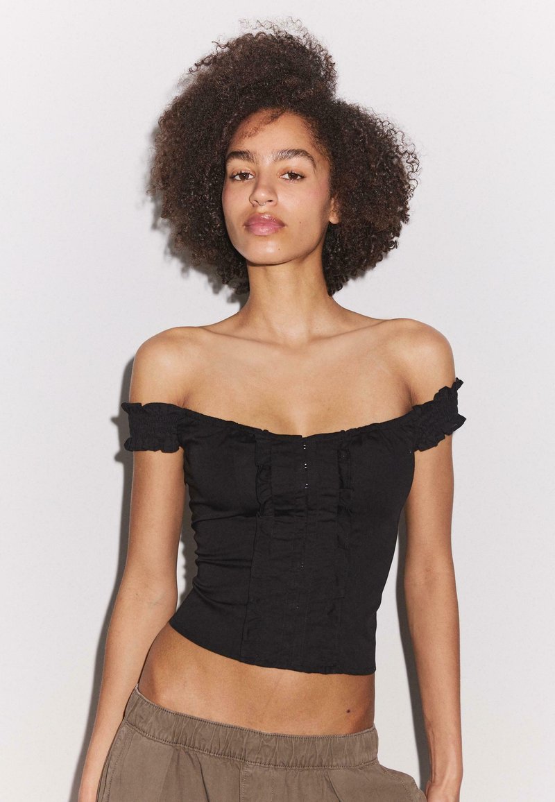Jeune femme aux cheveux bouclés portant un haut noir épaules dénudées et un pantalon marron, debout devant un fond clair uni.