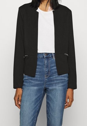Femme portant un blazer noir avec des détails zippés sur une chemise blanche et un jean bleu taille haute devant un fond blanc uni.