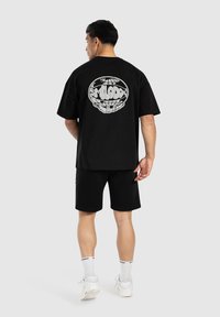 Camiseta negra de gran tamaño con diseño gráfico blanco en la parte de atrás, combinada con pantalones cortos negros, zapatillas blancas y calcetines estampados.