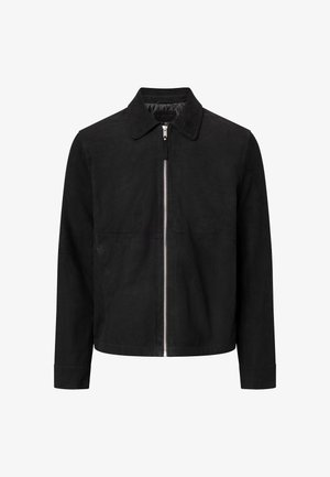 Junger Mann trägt eine schwarze Reißverschlussjacke über einem weißen Hemd und dunklen Hosen, schaut nach unten und hält am Reißverschluss der Jacke.