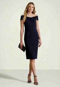 Mujer con un vestido negro de largo a la rodilla y sin hombros, sosteniendo un clutch negro, vistiendo sandalias de tacón alto negras, de pie frente a un fondo liso.