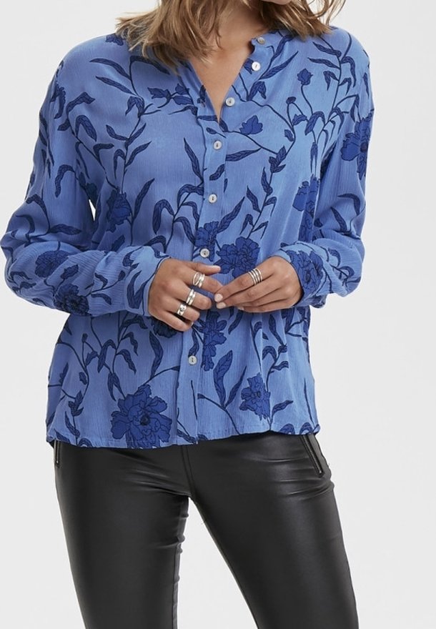 Femme portant un chemisier bleu à boutons avec des motifs floraux foncés et un pantalon en cuir noir, les mains jointes avec plusieurs bagues en argent.