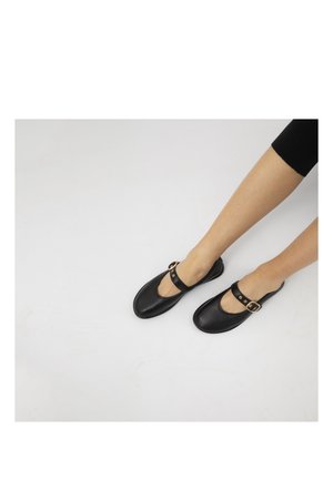 Beine in schwarzen, verkürzten Hosen, die schwarze Mary-Jane-Schuhe mit goldenen Schnallen auf einem hellgrauen Boden tragen.