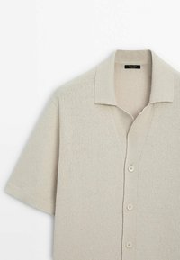 Vaaleanbeige neulottu lyhythihainen neuletakki, jossa on litteä kaulus ja viisi nappia. Pehmeä pinta, jossa on hienovarainen kudontamalli.