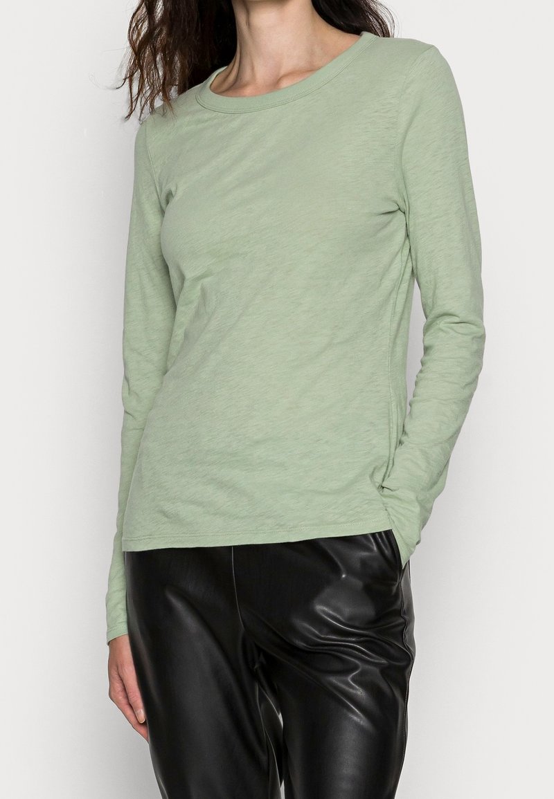Langärmliges, hellgrünes Oberteil aus weichem Material mit rundem Ausschnitt, kombiniert mit glänzenden, schwarzen Kunstlederhosen. Strukturiertes, tailliertes Design.