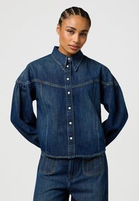 Veste en denim bleu foncé avec un devant à boutons, un col pointu et des manches volumineuses. Présente des coutures contrastées et un design court.