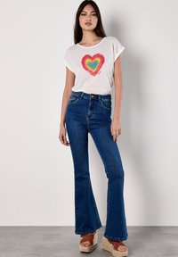 T-shirt blanc à manches courtes avec un graphique de cœur coloré, associé à un jean flare bleu taille haute et des sandales à plateformes beiges.