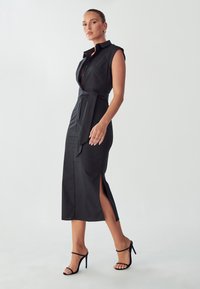 Vestido negro de camisa con cuello y cierre de botones en la parte delantera, incluye un cinturón de lazo, aberturas laterales y una textura suave y elegante. Llevado con tacones negros.