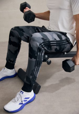 Szare dresowe spodnie w kamuflażu z czarnymi akcentami, dopasowanymi mankietami i trzema czarnymi paskami po boku. Białe buty sportowe z niebieskimi detalami.