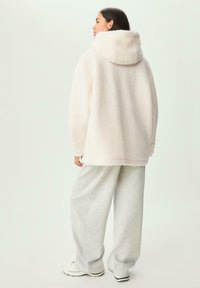 Femme portant une veste à capuche douce et rose clair, un pantalon gris ample et des baskets blanches, se tenant contre un fond neutre, de dos.