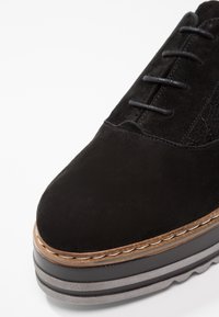 Chaussure à plateforme en daim noir avec lacets à l’avant, bordure marron cousue, et semelle superposée noire et grise.