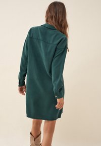 Vestito verde a maniche lunghe con colletto, realizzato in tessuto morbido. Presenta polsini con bottoni e orlo grezzo con spacco laterale. Design semplice e minimale.