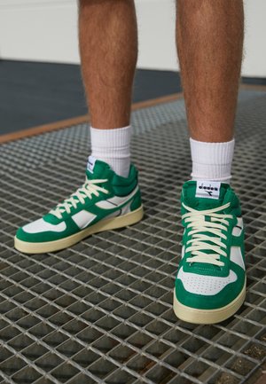 Personne portant des baskets Diadora vertes et blanches avec des lacets blancs et des chaussettes blanches, debout sur une surface en métal grillagée.