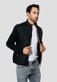 Čierna bomber bunda s prešívanou prednou časťou, oranžovým zipsom a rebrovanými manžetami. Nosená cez biele tričko so sivými džínsami.