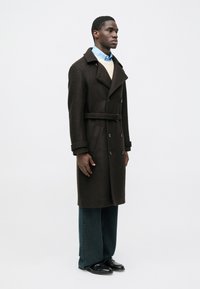 Abrigo cruzado de mezcla de lana marrón con grandes solapas, cintura con cinturón, bolsillos laterales, llevado sobre un suéter de color claro y una camisa azul.