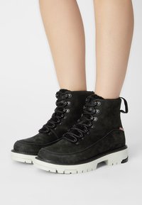 Botas de tobillo de ante negro con un acabado texturizado, parte frontal con cordones, suela de goma resistente y tirador trasero. Acentuadas con ojales de metal y detalle del logo.