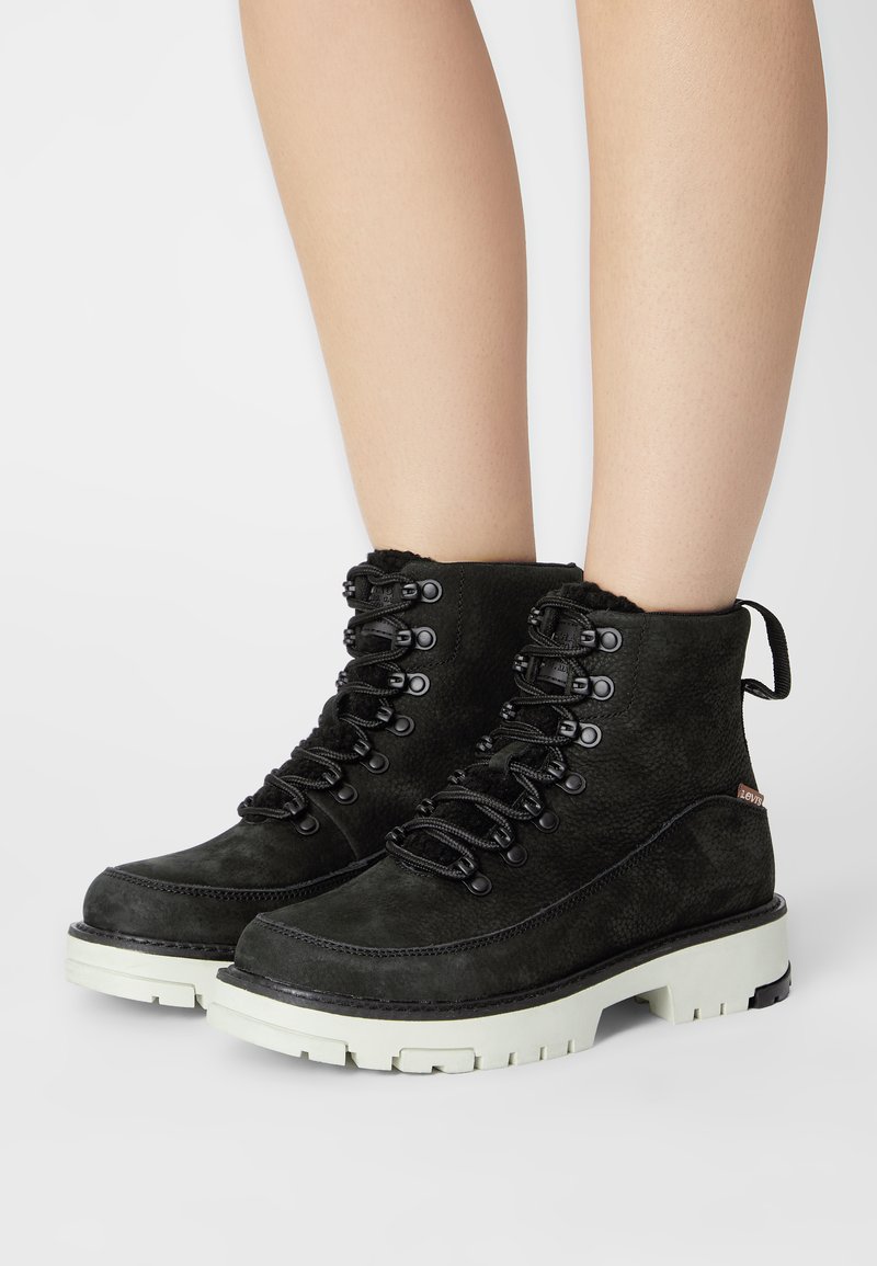 Botas de tobillo de ante negro con un acabado texturizado, parte frontal con cordones, suela de goma resistente y tirador trasero. Acentuadas con ojales de metal y detalle del logo.