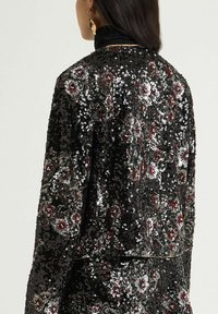 Veste noire à sequins avec motifs floraux rouges et argentés. Présente une surface texturée et des détails complexes. Silhouette élégante.