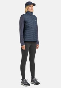 Marineblaues wattiertes Westchen mit hohem Kragen, schwarzes Langarmoberteil, schwarze Leggings und graue Trailschuhe, kombiniert mit einer passenden Mütze.
