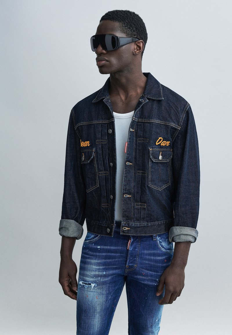 Hombre con gafas de sol negras grandes, chaqueta de mezclilla oscura con mangas enrolladas, camiseta blanca sin mangas y jeans azules desgastados contra un fondo gris.