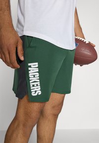Zaļas sporta šortus ar sānu paneļiem tumši pelēkā krāsā, kuru priekšpusē ir uzraksts "PACKERS" ar treknajiem baltiem burtiem. Izgatavoti no viegla, gluda materiāla.