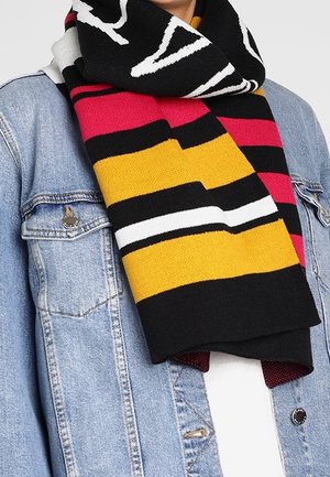 Personne portant une écharpe multicolore à rayures noires, jaunes, blanches et rouges par-dessus une veste en jean bleu clair et une chemise blanche.