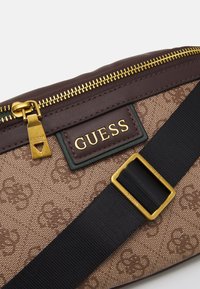 Brun mönstrad canvasväska med en gyllene dragkedja, GUESS-logotyp i guld på ett mörkbrunt fält, justerbar svart rem med detaljer i guld.