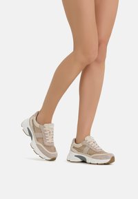 Beige athletic shoes with mesh upper, textured accents, and white sole. Features delicate laces and metallic details on the sides.