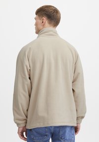 Beige fleece jacket with a high collar and long sleeves. Features a relaxed fit and textured, soft surface. No visible logos or patterns.