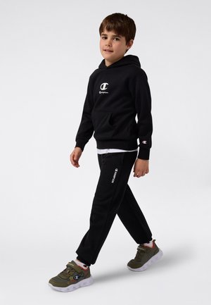 Ragazzo che cammina indossando un hoodie nero Champion, pantaloni sportivi neri, calzini bianchi e sneakers verdi con suole bianche su sfondo bianco.
