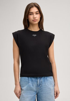 Kobieta o prostych brązowych włosach, ubrana w czarny, bezrękawnik z poduszkami na ramionach z logo "JOOP! JEANS" oraz jasnoniebieskie dżinsy, na jednolitym tle.