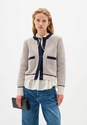 Kreminė megzta cardigan su tamsiai mėlynomis detalėmis, apvaliu kaklu, dviem priekinėmis kišenėmis, sagų uždarymu ir banguotais rankogaliais. Dėvima ant permatomo marškinėlio.